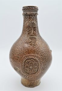 A brown jug with a mottled pattern, featuring a face and a rose seal in the centre