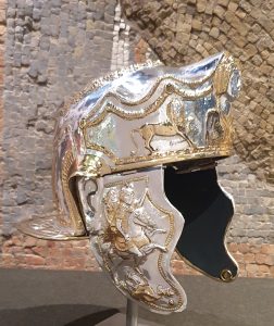 A shining silver and gold Roman cavalry helmet sits on a stand in front of a mosaic.