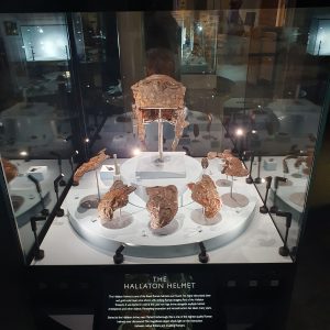 A display case containing a corroded Roman cavalry helmet on an elevated stand, surrounded by five cheekpieces, with a text label in front of the case.