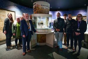 A group of people stand around a circular museum display, which contains a shining replica Roman cavalry helmet.