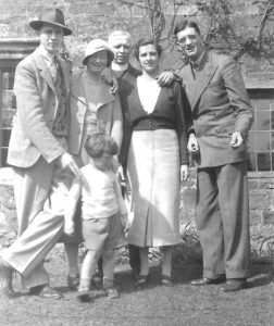 Photograph of five well-dressed adults and one child in the sunshine in front of an old house
