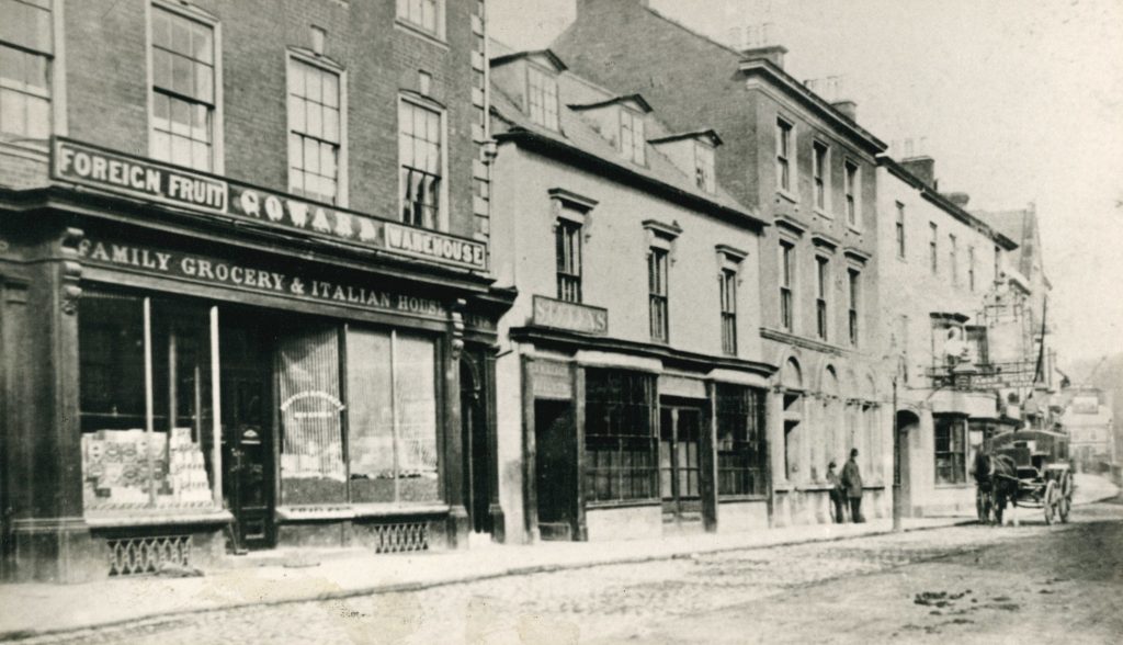 An old photograph of shops including a grocers, with a horse and carriage passing on the road