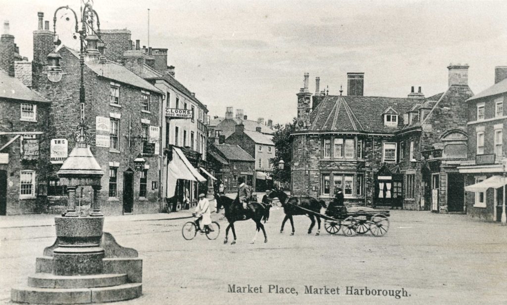 An old photo of a market place, with water pump in the foreground and horses and bicycles crossing the square