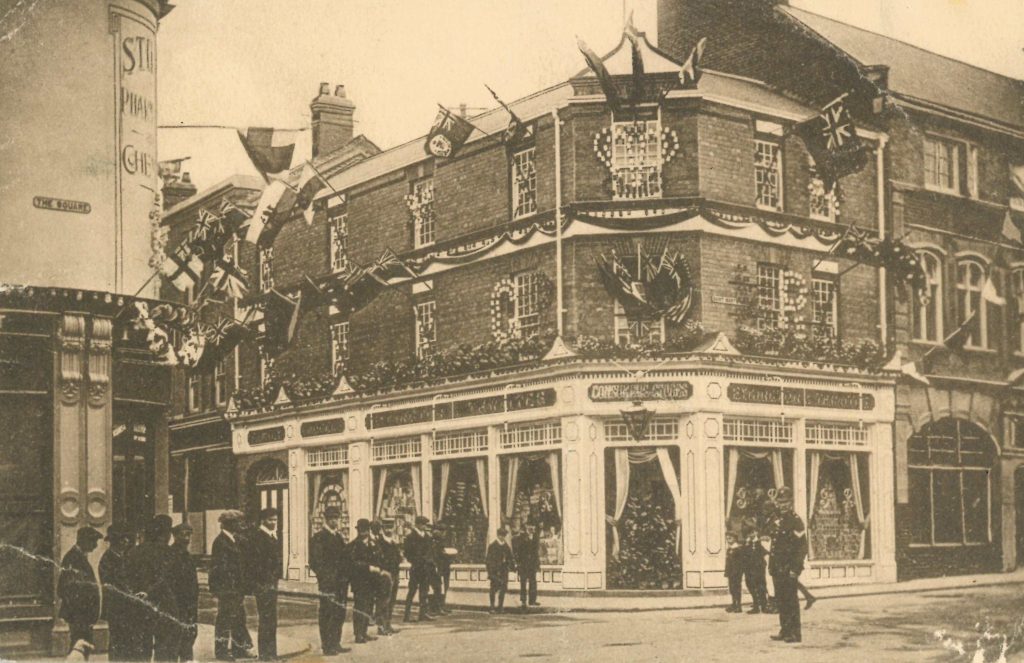 An old photograph of a grocery shop on a street corner, with frags hanging from the building and a row of men standing outside