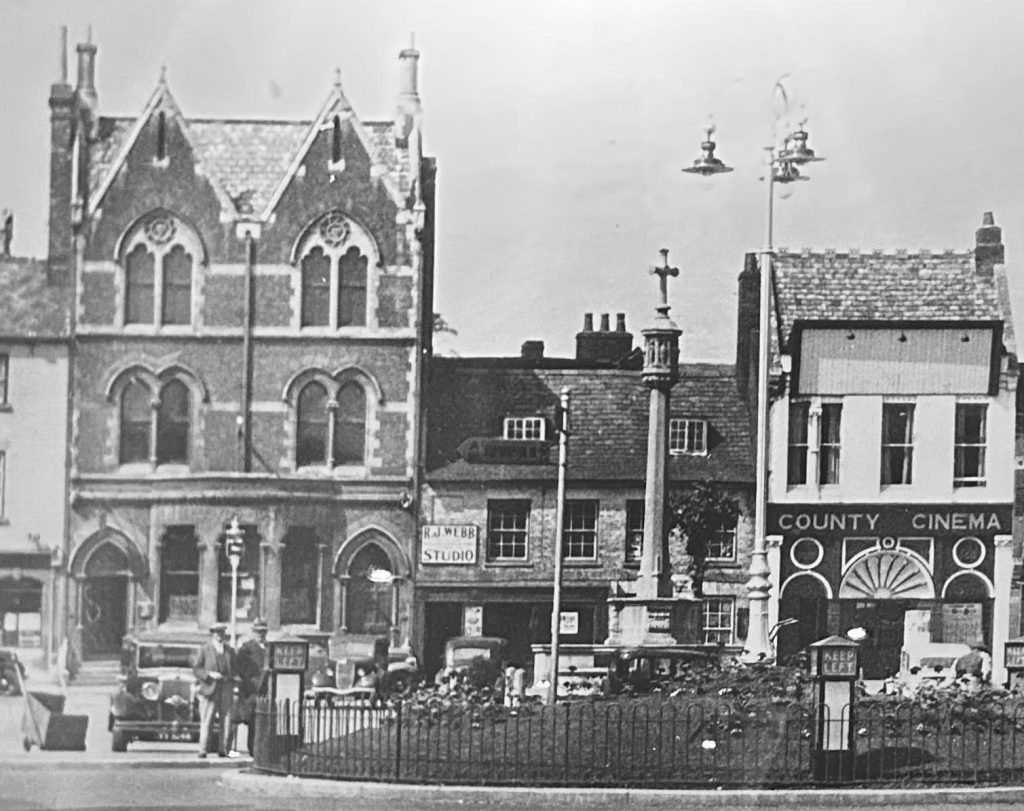 An old photograph of a row of shops and a cinema with a war memorial in the foreground